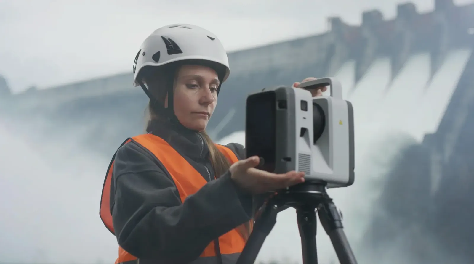Kvinne med hjelm og refleksvest stiller inn en laserskanner på stativ foran en demning